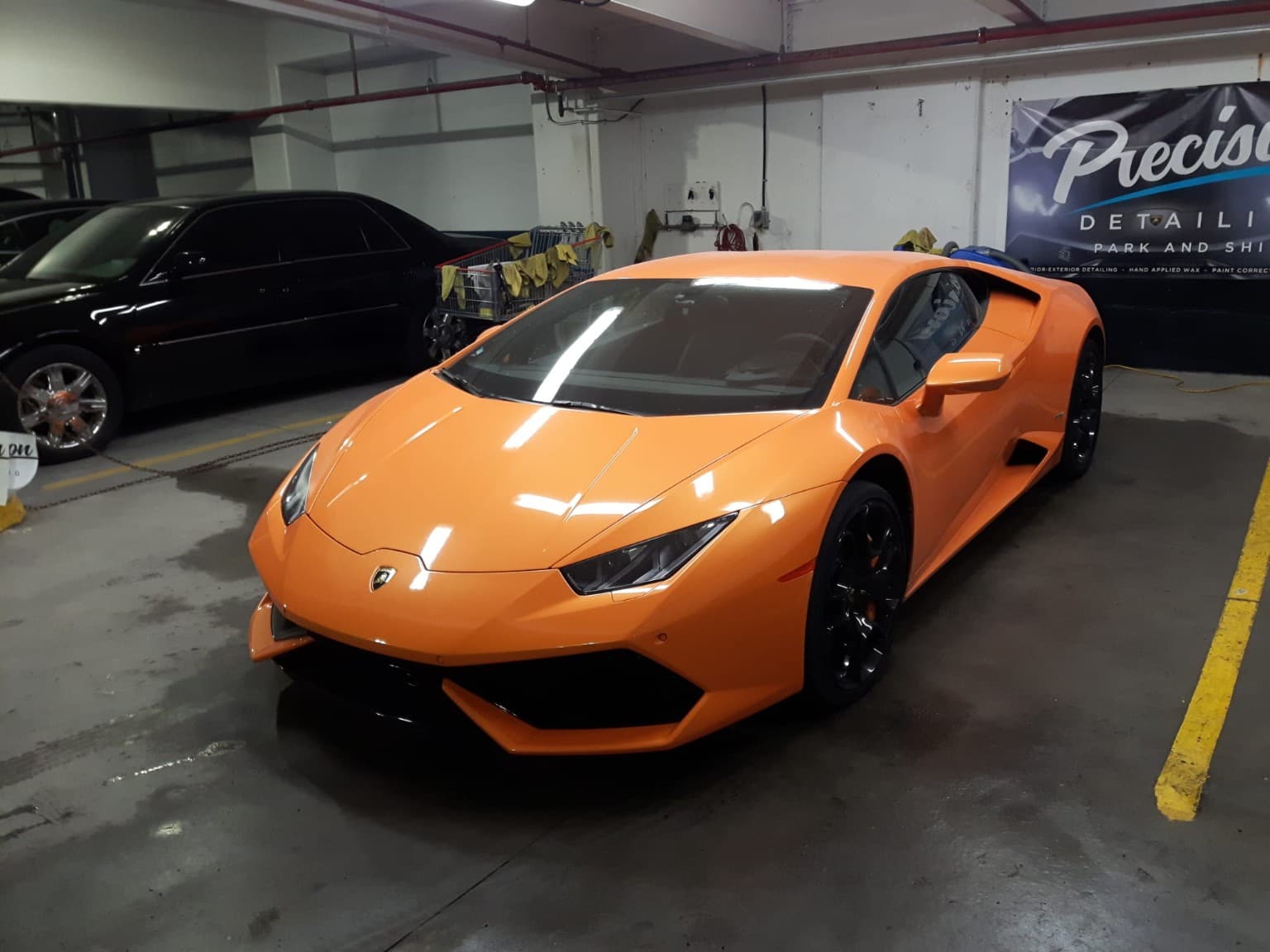 Vibrant orange Lamborghini Huracán parked in a detailing shop beside a black luxury sedan.