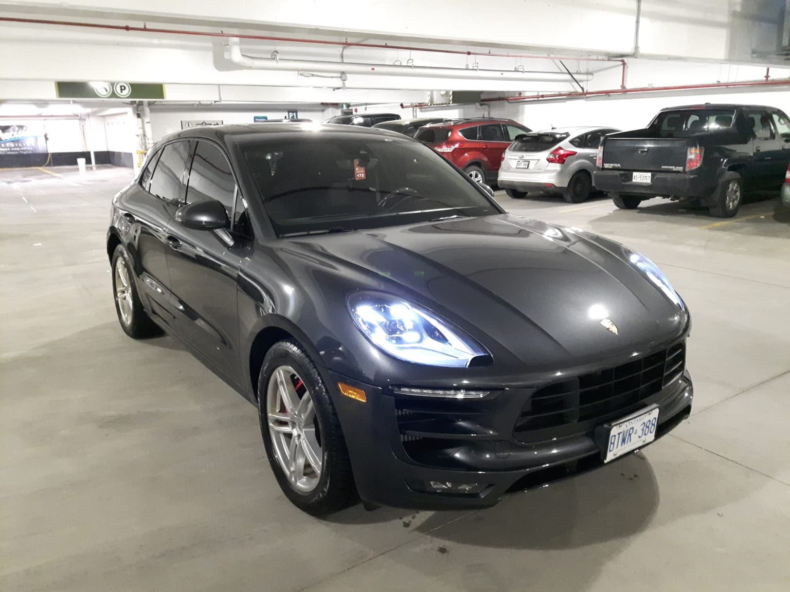 Dark grey Porsche Macan SUV with illuminated headlights parked in an indoor garage.